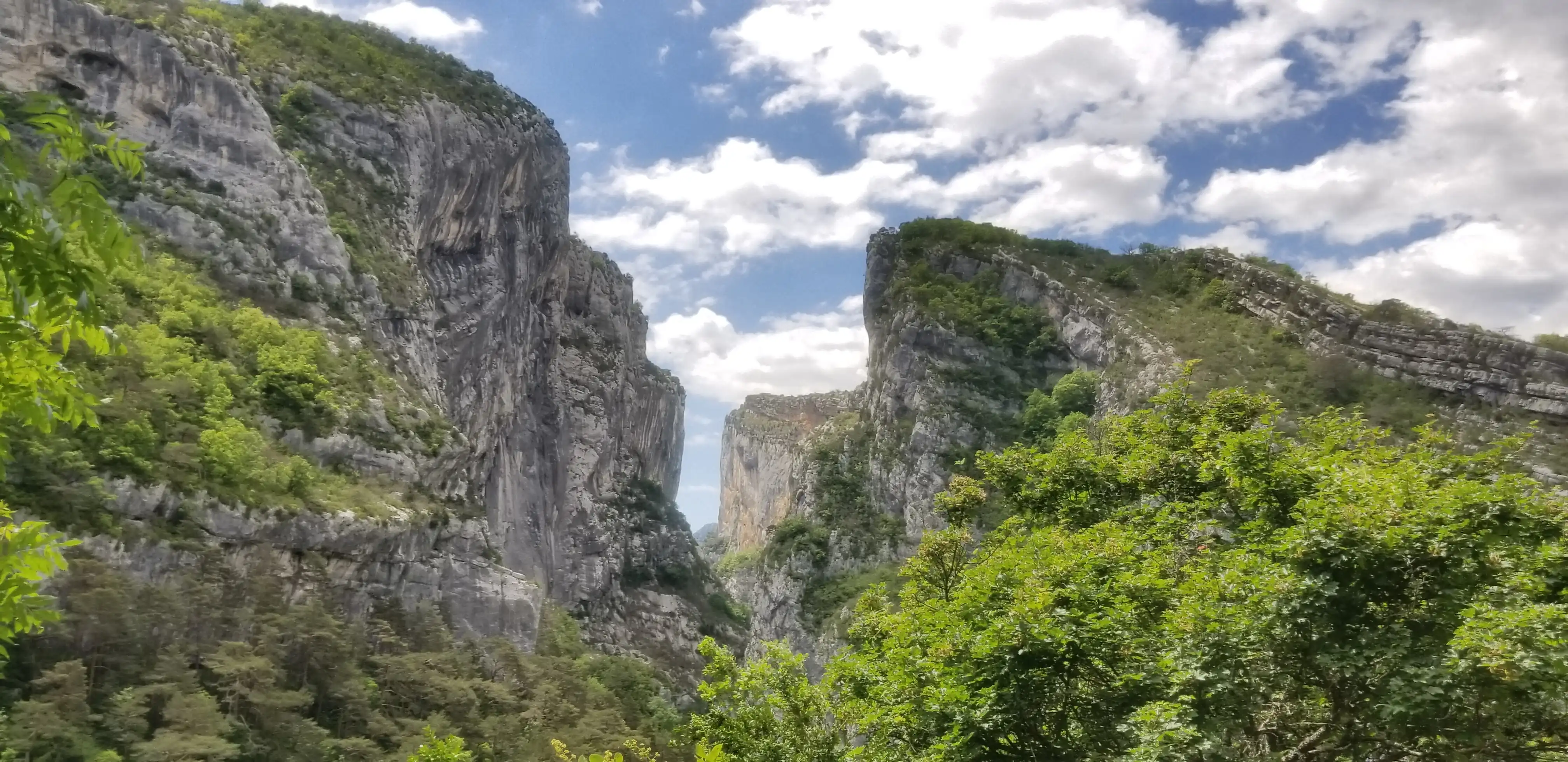 A dramatic mountain landscape showcasing towering rocky cliffs with lush greenery at their bases. The cliffs form a narrow passageway, creating a striking contrast against the vibrant blue sky filled with fluffy clouds. The dense vegetation in the foreground adds depth to this scenic view.